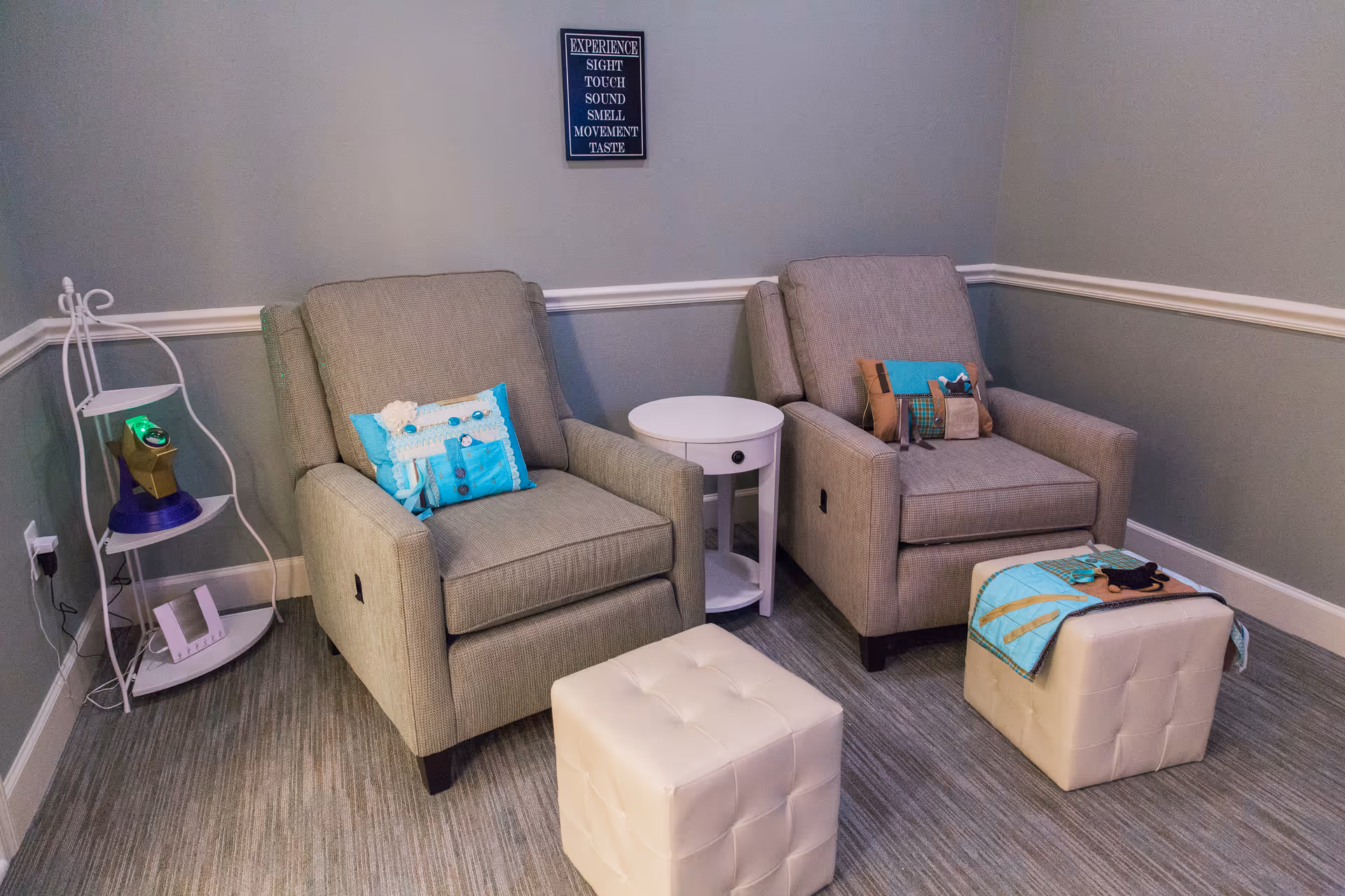 A cozy corner of a room with two gray upholstered armchairs, each with a decorative pillow. Between the chairs is a small round white side table. In front of each chair is a white tufted ottoman, one with a folded blue blanket on top. To the left is a white corner shelf with a few items including a small electronic device. The walls are painted gray with white trim, and a framed sign hangs above the chairs with the words: EXPERIENCE SIGHT TOUCH SOUND SMELL MOVEMENT TASTE.