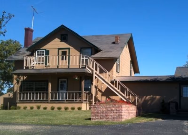 A two-story beige house with a front porch and an external staircase leading to a second-floor balcony. The house has a chimney, several windows, and a well-maintained lawn with small shrubs. The sky is clear and blue.
