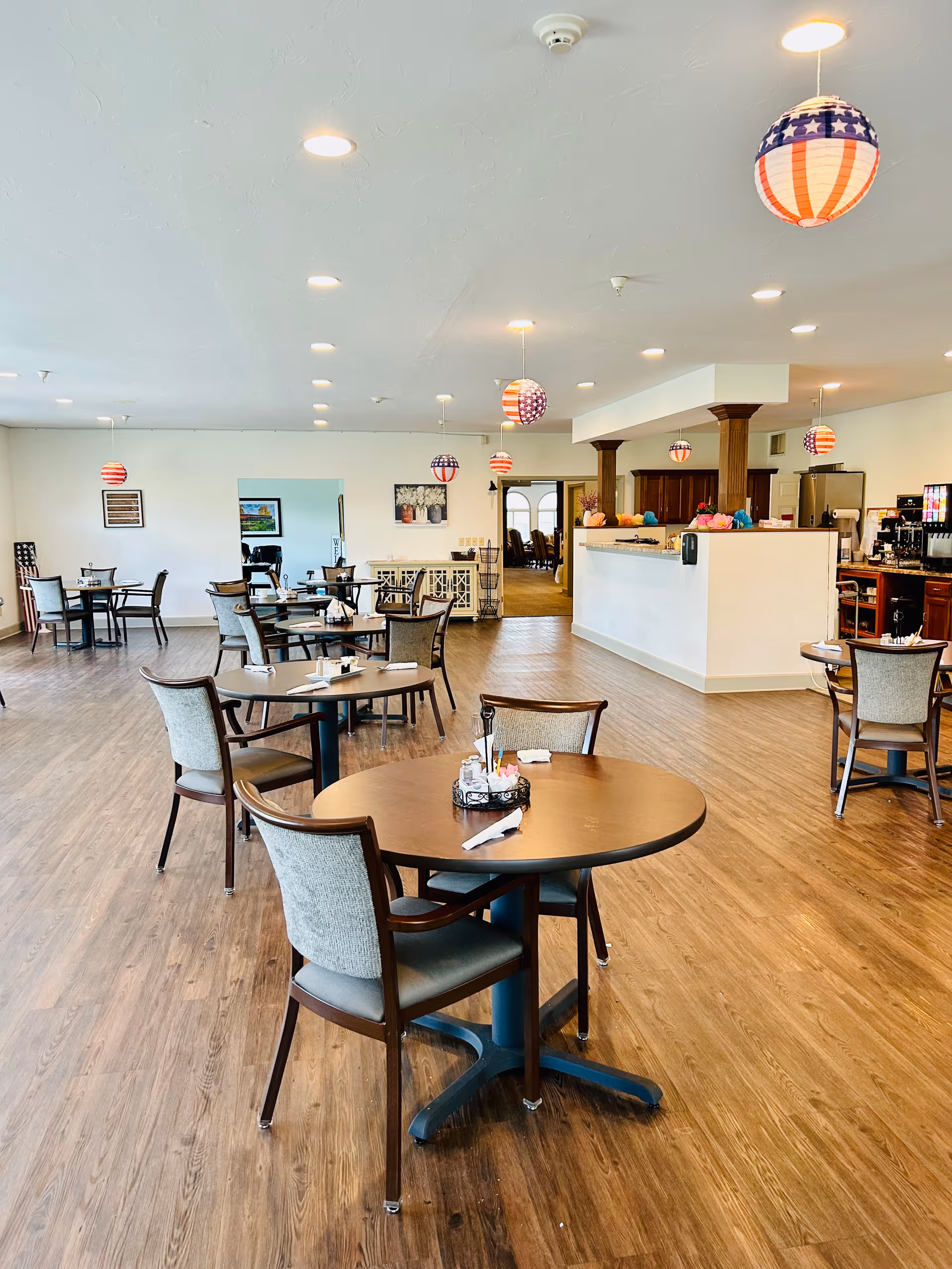 Bright dining room with round tables and chairs, a service counter, and hanging patriotic lanterns.