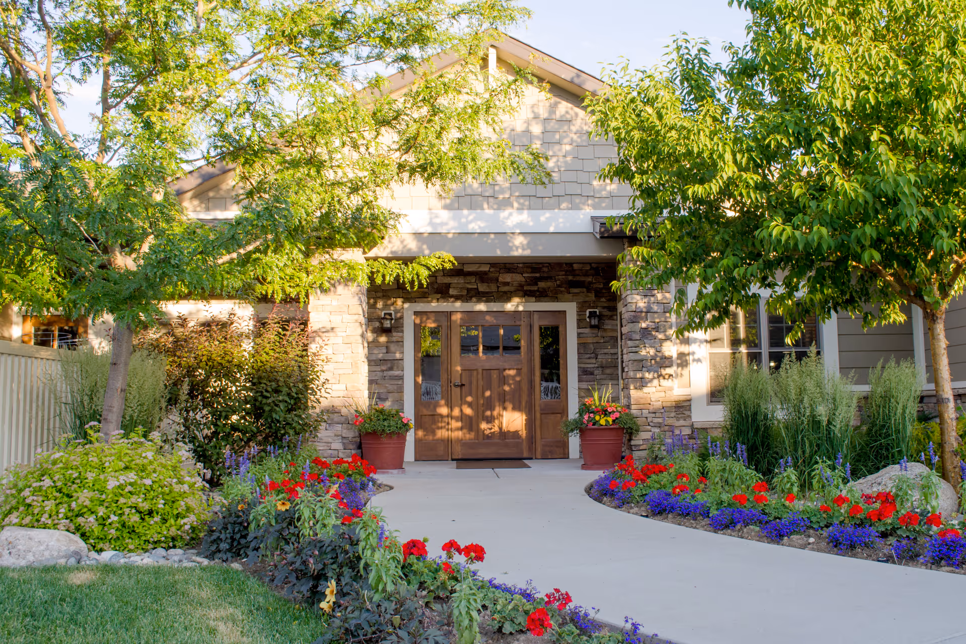 Front entrance of a building with a stone facade and wooden double doors, surrounded by lush greenery and colorful flower beds along a curved concrete pathway.