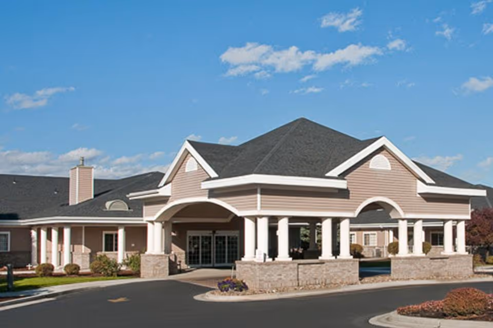 Front exterior view of a single-story building with a covered entrance supported by white columns, beige siding, and a dark gray roof under a blue sky with scattered clouds.