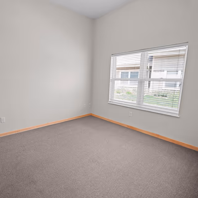 Empty room with beige walls, a window with white blinds showing an outside view of another building, and a carpeted floor with wooden baseboards.