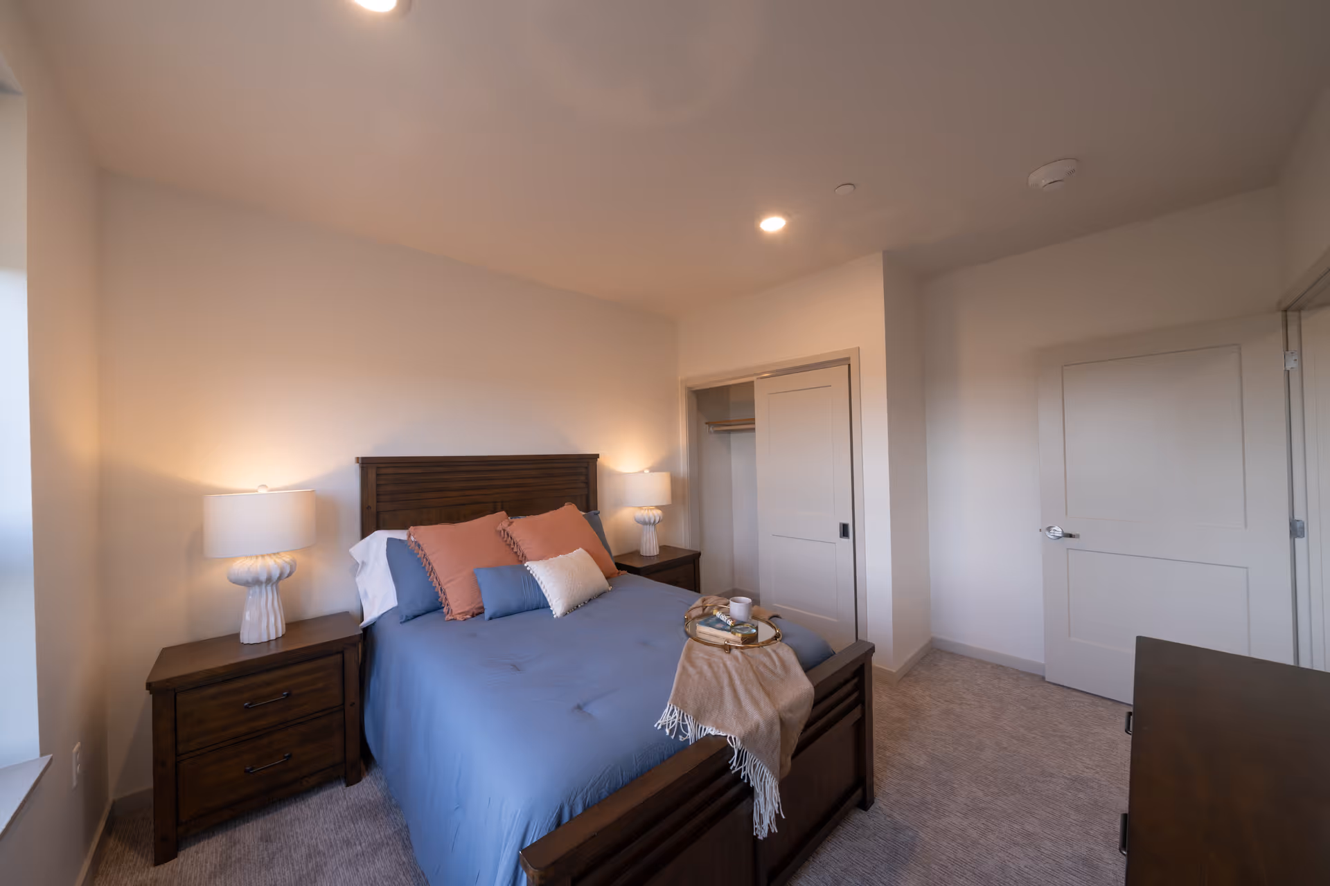 A cozy bedroom with a wooden bed frame and headboard, dressed in blue bedding with several pillows in shades of blue, white, and peach. Two matching wooden nightstands flank the bed, each with a white lamp. There is a small tray with a cup and a book on the bed. The room has beige walls, a carpeted floor, a closet with sliding doors, and a closed door leading out of the room.