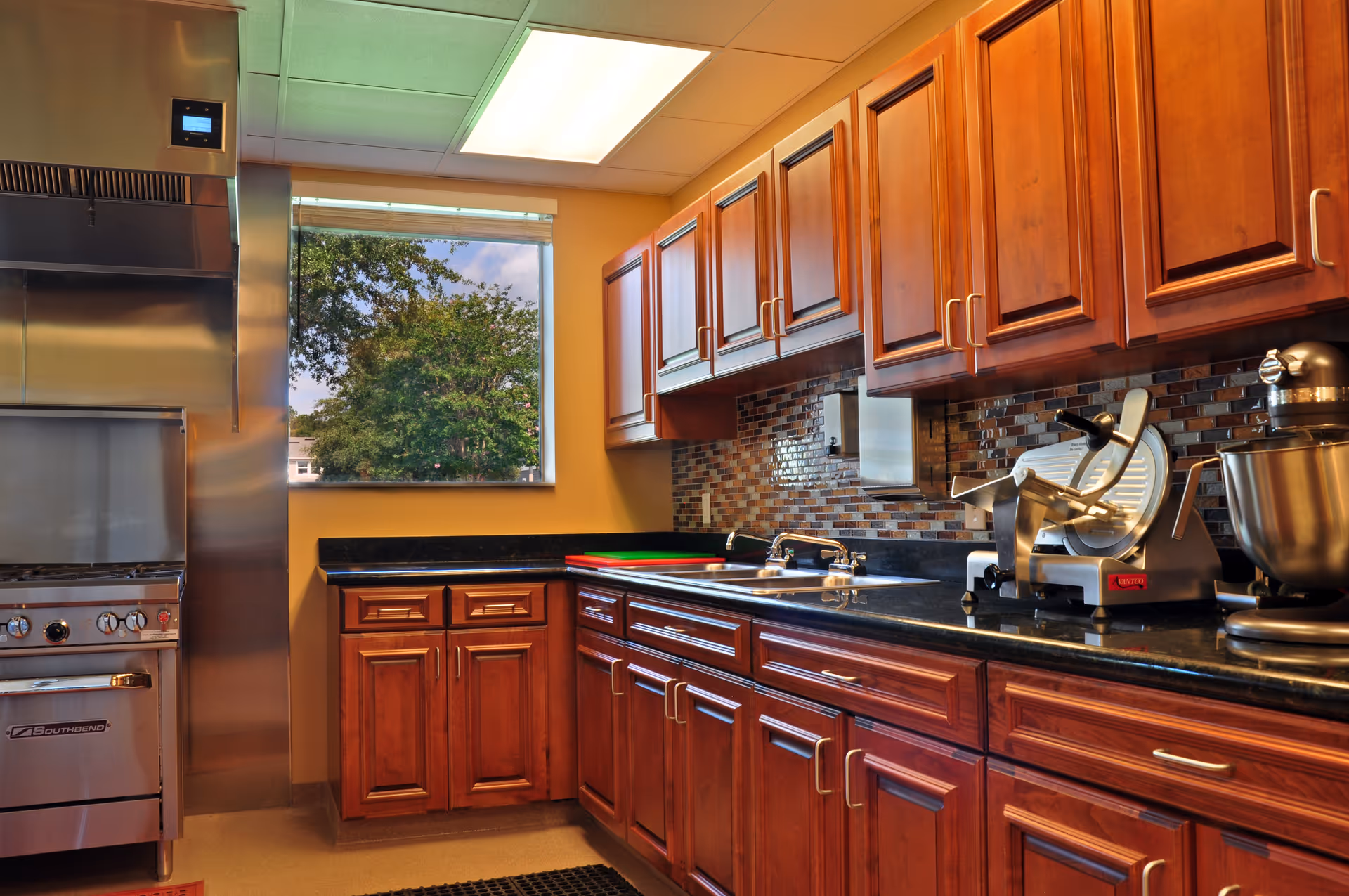 A kitchen with wooden cabinets, a black countertop, a stainless steel stove and oven, a window showing trees outside, a sink, a meat slicer, and a stand mixer.