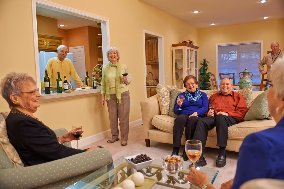 A group of elderly people socializing in a cozy living room. Four seniors are seated on sofas and chairs holding glasses of wine, while two others stand near a kitchen pass-through counter with bottles of wine. The room is warmly lit with beige walls and decorated with plants and a cabinet.
