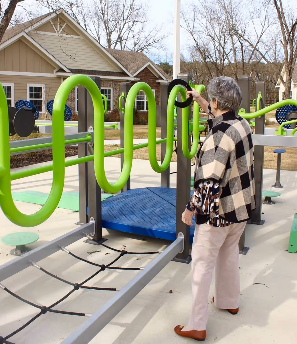 An elderly woman wearing a checkered shawl and light-colored pants is standing outdoors at a senior living facility playground or exercise area. She is interacting with bright green and blue exercise equipment designed for seniors. In the background, there are residential-style buildings and leafless trees under a partly cloudy sky.