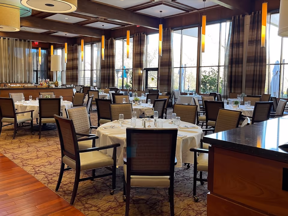 Spacious dining room with round tables set with white tablecloths and chairs, large windows and hanging pendant lights.
