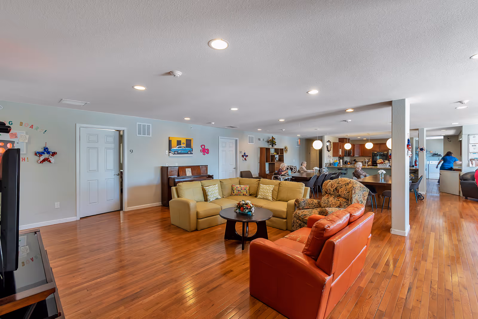 A spacious senior living common area with wooden flooring, featuring a yellow sectional sofa, a floral armchair, and a red leather armchair around a round coffee table. In the background, there are dining tables with chairs and a kitchen area with pendant lights. Several seniors are seated at the tables, and a staff member is visible near the kitchen. The walls are decorated with colorful artwork and a piano is against one wall.