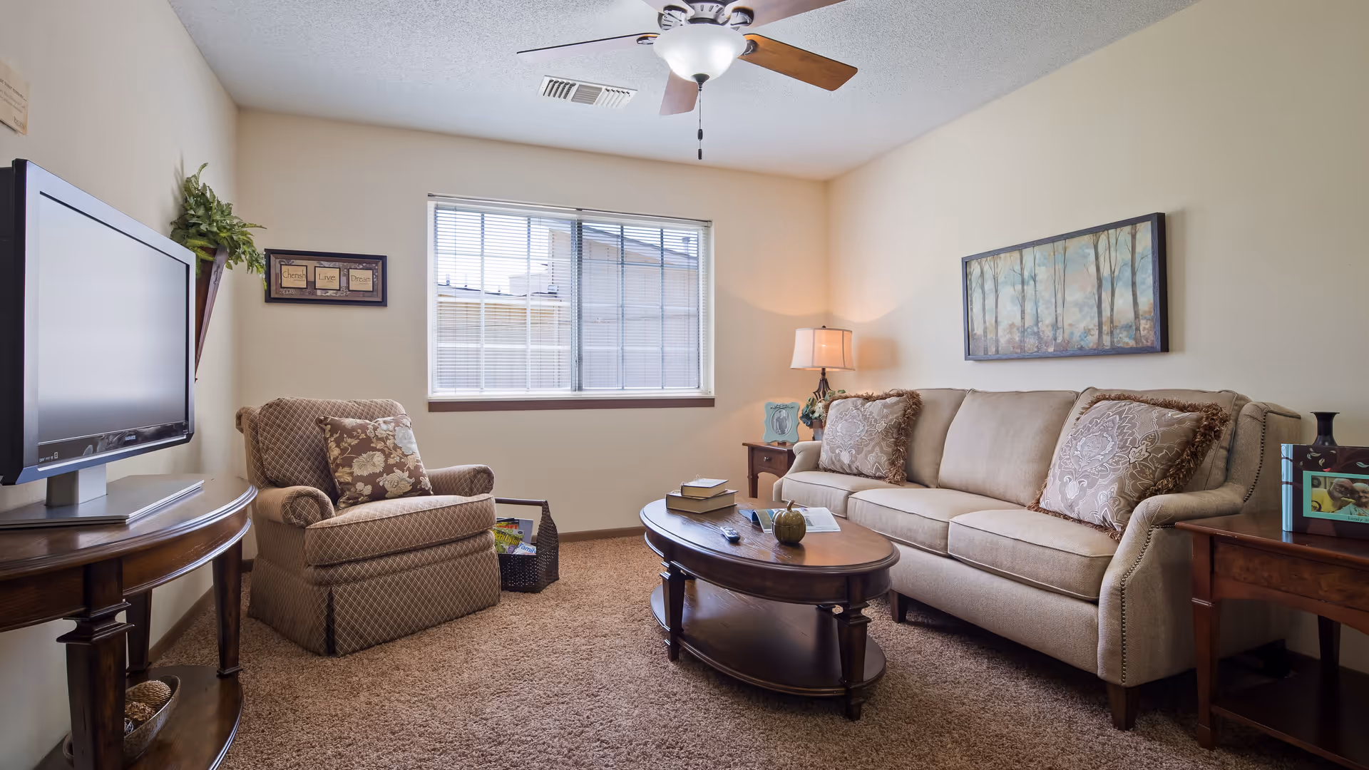 A cozy living room with a beige sofa adorned with patterned pillows, a matching armchair, a wooden coffee table with books and a decorative item, a flat-screen TV on a wooden stand, a window with blinds, a ceiling fan with light, and framed artwork on the walls.