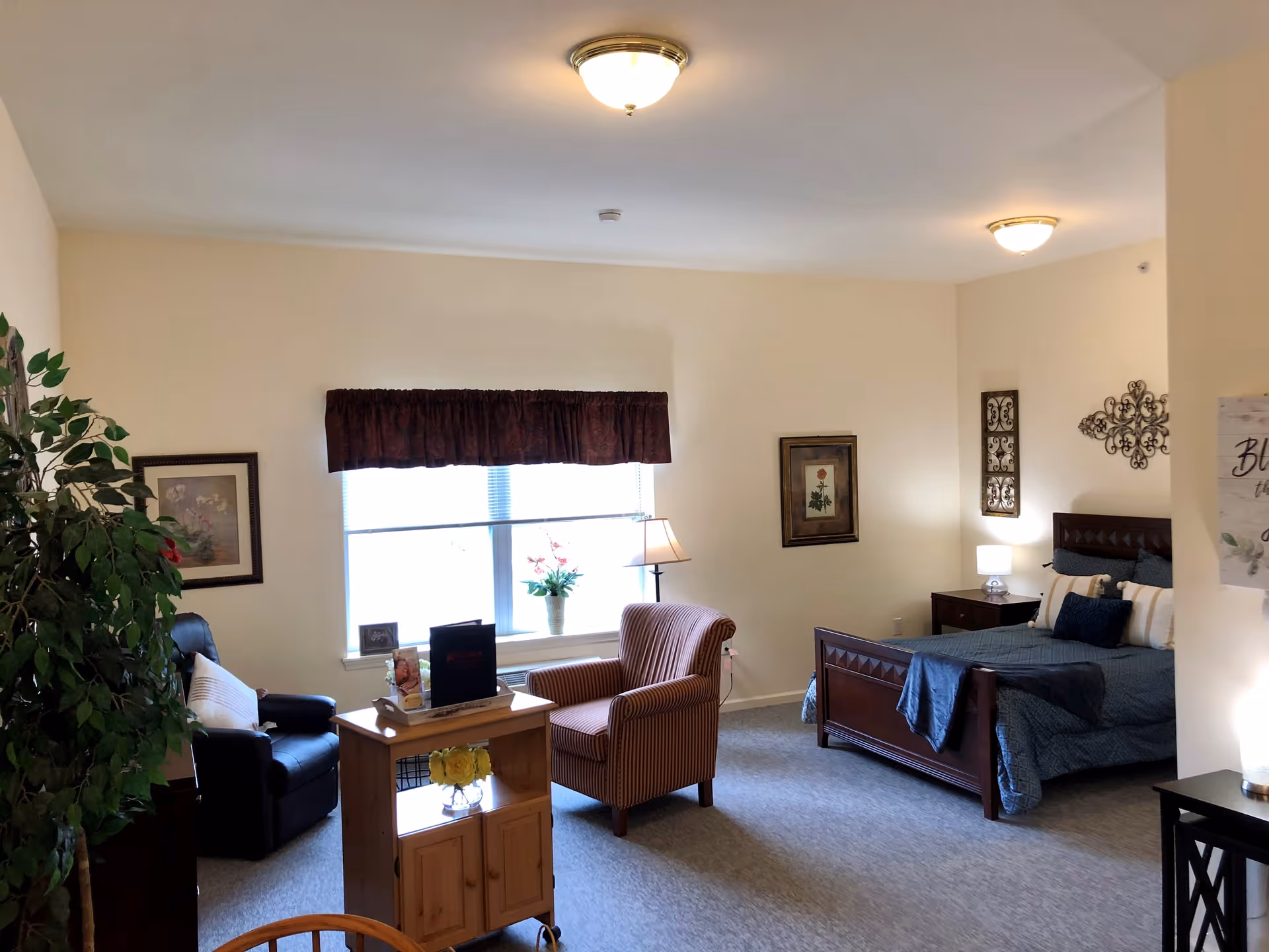 A cozy bedroom in a senior living facility with a wooden bed dressed in blue bedding, a striped armchair, a black recliner, a wooden cabinet with flowers, framed artwork on the walls, and a window with a dark valance letting in natural light.