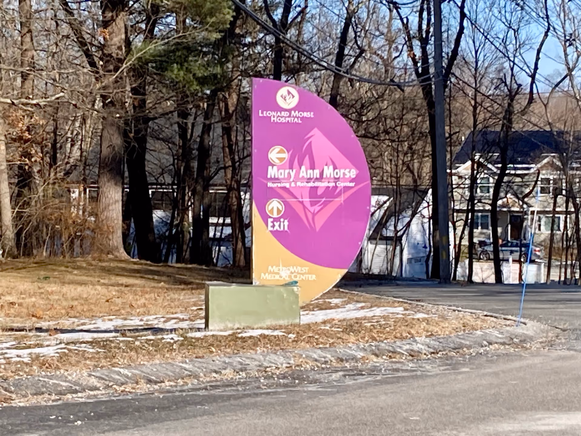 Outdoor scene showing a purple and yellow directional sign for Leonard Morse Hospital and Mary Ann Morse Nursing & Rehabilitation Center, with arrows indicating directions. The sign is situated on a grassy area near a road, with leafless trees and houses in the background.