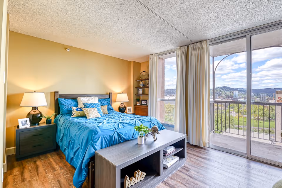 A bright bedroom with a large bed covered in a blue comforter and multiple decorative pillows. There are two bedside tables with lamps and framed photos. A small shelving unit with decorative items is in the corner. The room has wood flooring and large sliding glass doors leading to a balcony with a view of a cityscape and hills under a partly cloudy sky.