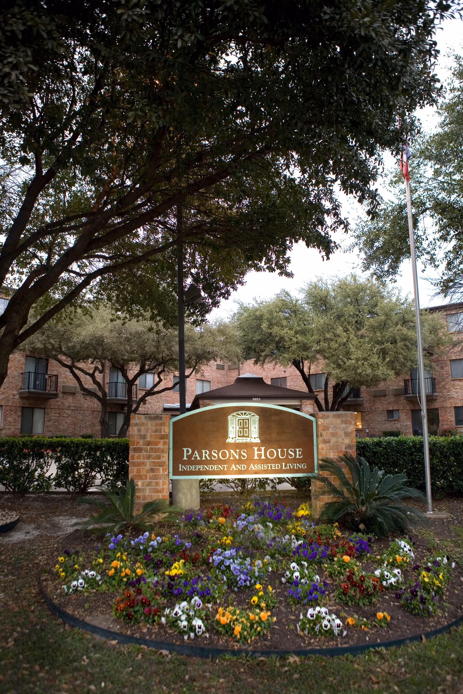 Outdoor view of Parsons House Independent and Assisted Living facility sign surrounded by colorful flowers, with trees and a brick building in the background.