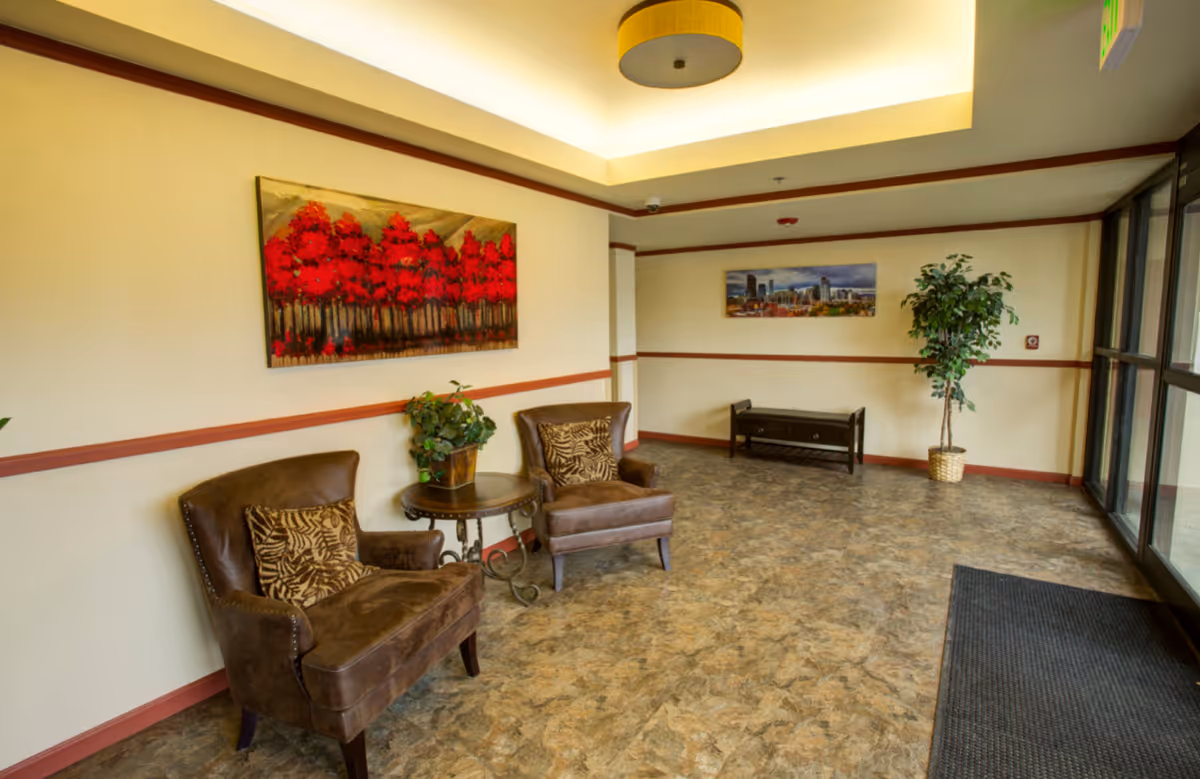 A waiting area with two brown leather armchairs with patterned cushions, a small round table with a plant, a bench against the far wall, two framed pictures on the walls, a potted plant, and large glass doors letting in natural light.