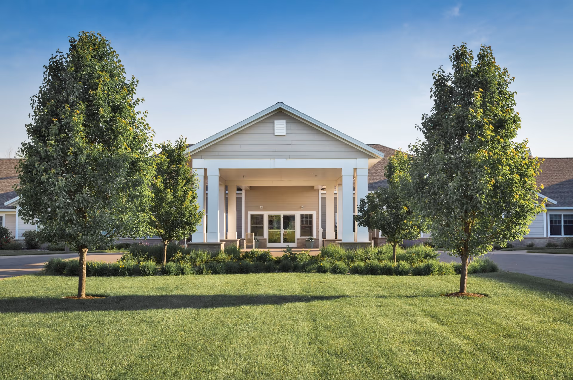 Front entrance of a senior living community building with a covered portico, manicured lawn, and trees.
