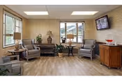 A cozy sitting area with four gray armchairs arranged around two small round tables, a wooden cabinet with a red decorative item on top, a wall-mounted flat-screen TV, and large windows letting in natural light. The room has light wood flooring and patterned wallpaper.