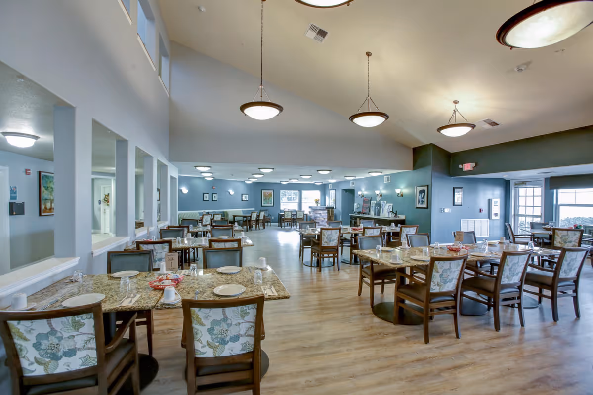 Spacious dining room with multiple set tables and floral-upholstered chairs in a senior living facility.