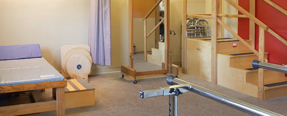 A rehabilitation room with a padded therapy bed, wooden stairs with handrails, parallel bars for walking exercises, a large mirror, and exercise equipment. The room has beige walls and carpeted flooring.