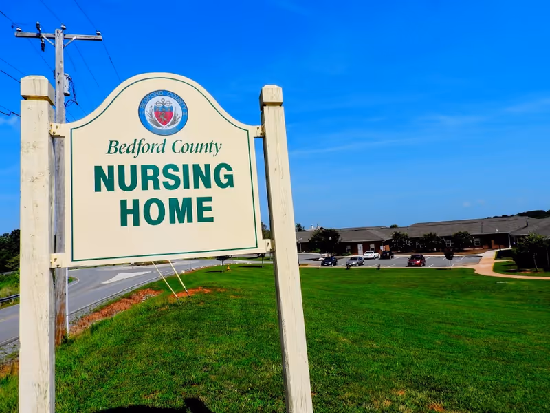 A large sign reading 'Bedford County Nursing Home' stands on a grassy area near a road. In the background, there is a single-story building with a parking lot containing several cars under a clear blue sky.