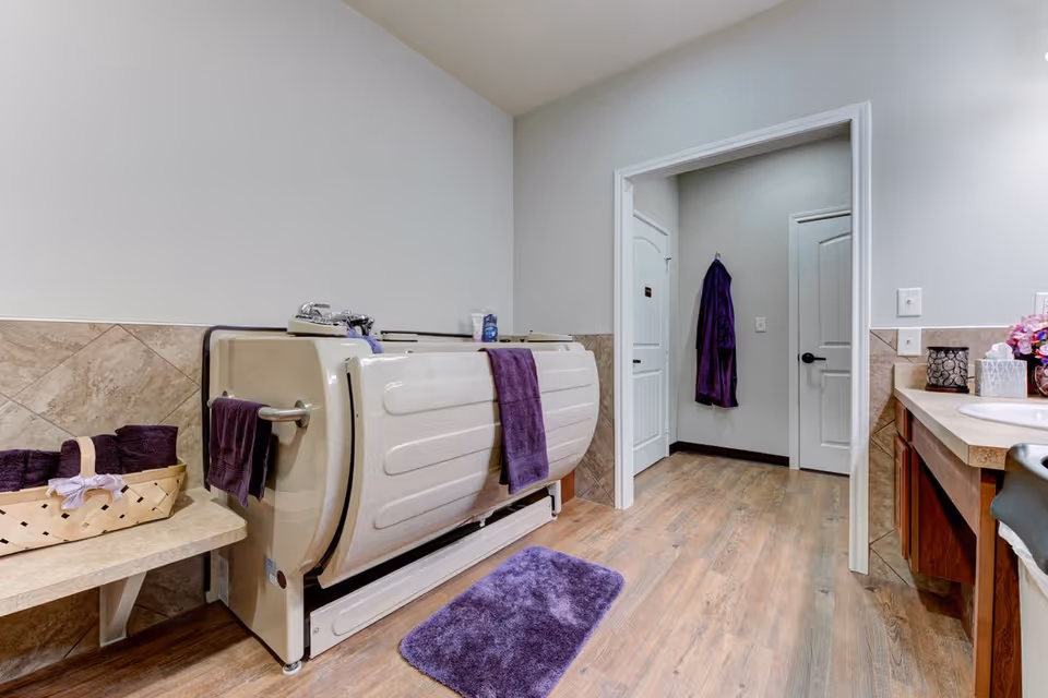 A senior living facility bathroom featuring a walk-in bathtub with a purple towel hanging on its side. The room has beige tiled walls, wooden flooring, a purple bath mat, and a basket with purple towels on a small shelf. There is a vanity with a sink and decorative items on the right side, and a purple robe hanging on the wall in the hallway visible through an open doorway.