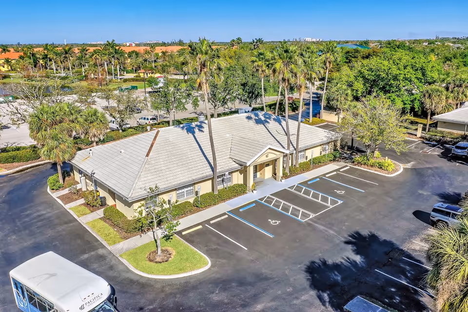 Aerial view of a single-story building with a light gray roof surrounded by palm trees and greenery. The building is situated in a parking lot with several marked parking spaces, including handicapped spots. There is a white shuttle bus parked in the lot, and the surrounding area features more trees and a clear blue sky.