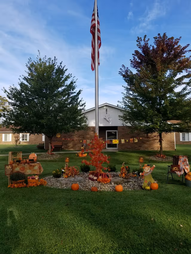 Outdoor scene at LECOM at Snyder Memorial featuring a flagpole with an American flag, two large trees, and a festive autumn display with pumpkins, fall decorations, and a scarecrow on a grassy lawn in front of a brick building.