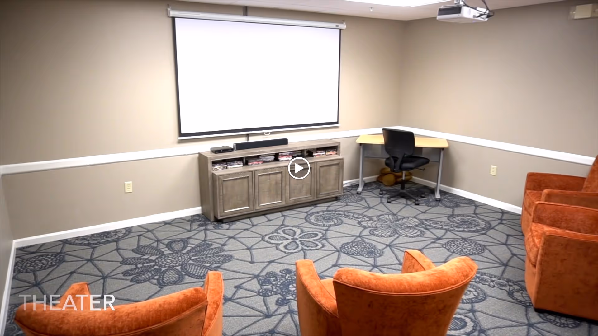 A small theater room with a patterned carpet, four orange upholstered chairs arranged facing a large white projection screen mounted on a beige wall. Below the screen is a wooden cabinet with media equipment and DVDs. A corner desk with a black office chair is positioned to the right side of the room.