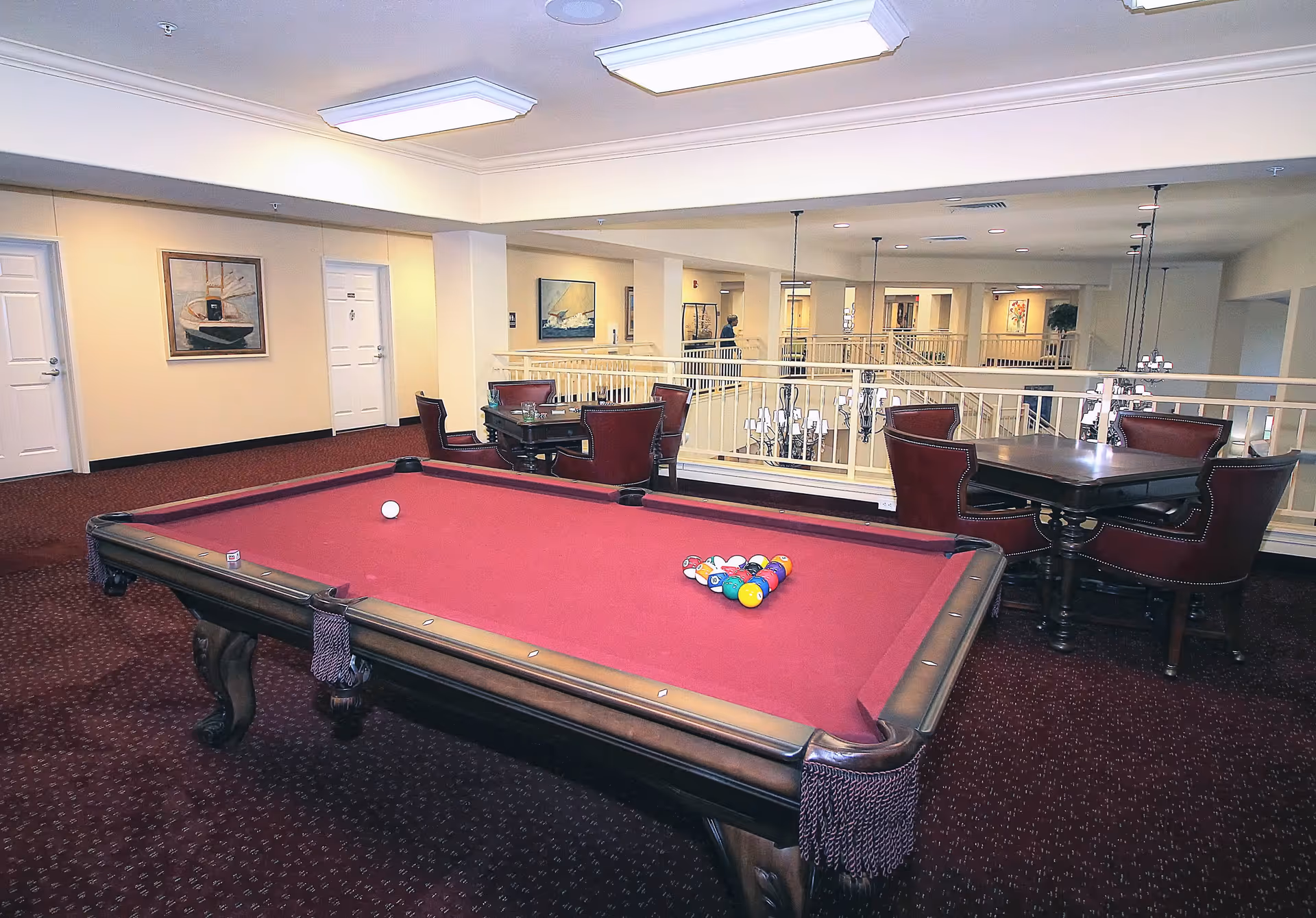 Interior view of a recreational area in a senior living facility featuring a red felt pool table with balls arranged for a game. Behind the pool table, there are two tables with four red leather chairs each. The area is carpeted in dark red and has beige walls with framed artwork. The space overlooks a lower level with railings and hanging light fixtures.