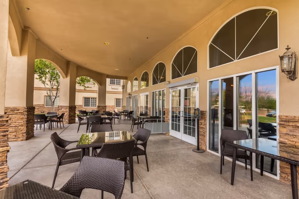 Outdoor covered patio area with multiple tables and chairs arranged for seating. The patio has stone pillars and large windows and doors leading inside the building. Trees and greenery are visible outside.