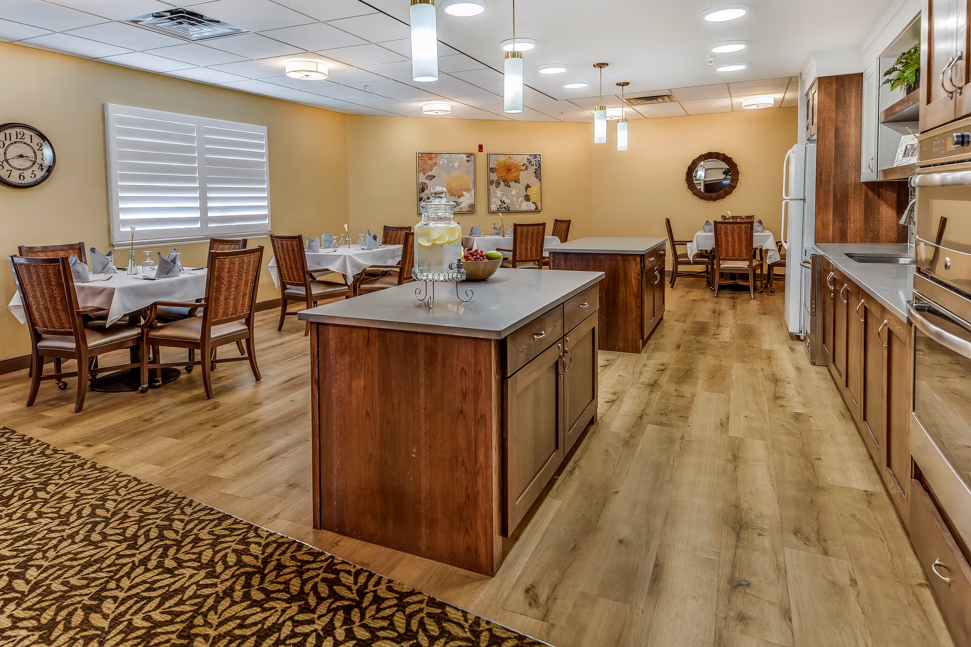 A dining area in a senior living facility featuring multiple tables with chairs, each table set with white tablecloths, napkins, and small vases with flowers. The room has wooden flooring and two kitchen islands with a water dispenser and a bowl of fruit on one. The kitchen area includes cabinets, a refrigerator, and built-in ovens. The walls are decorated with framed floral artwork and a round mirror.