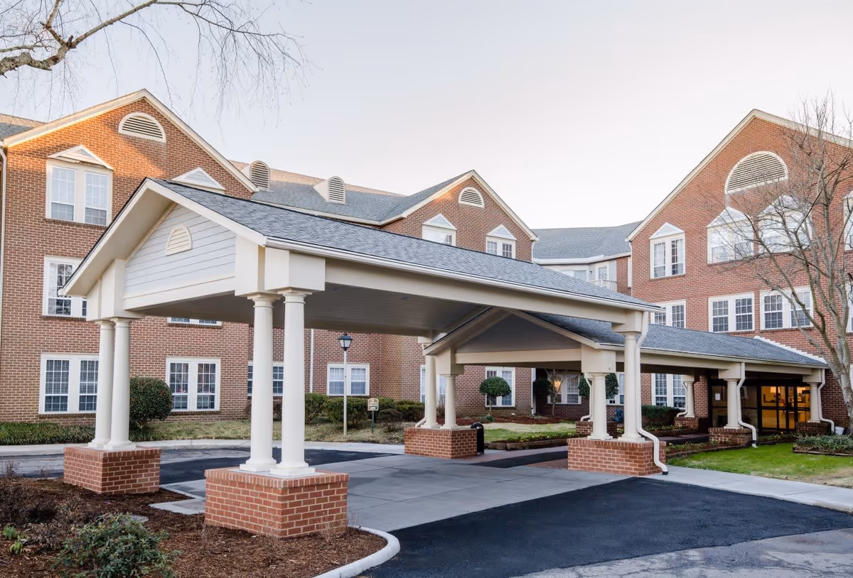 Exterior view of a senior living facility named First Colonial Inn featuring a large covered entrance supported by white columns with brick bases. The building is made of red brick with multiple windows and a gray shingled roof. There are some bushes and trees around the entrance area.