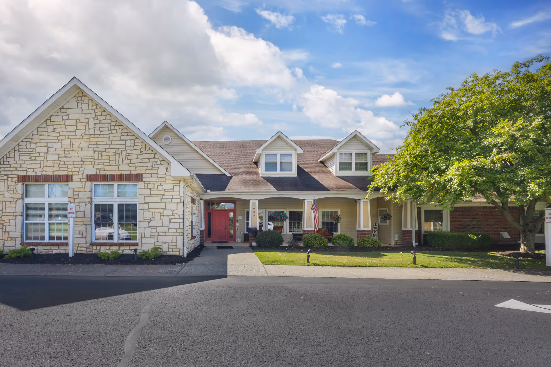 Front exterior view of a single-story senior living facility building with a stone and siding facade, a red entrance door, several windows, a small lawn with bushes, and a large tree on the right under a partly cloudy sky.