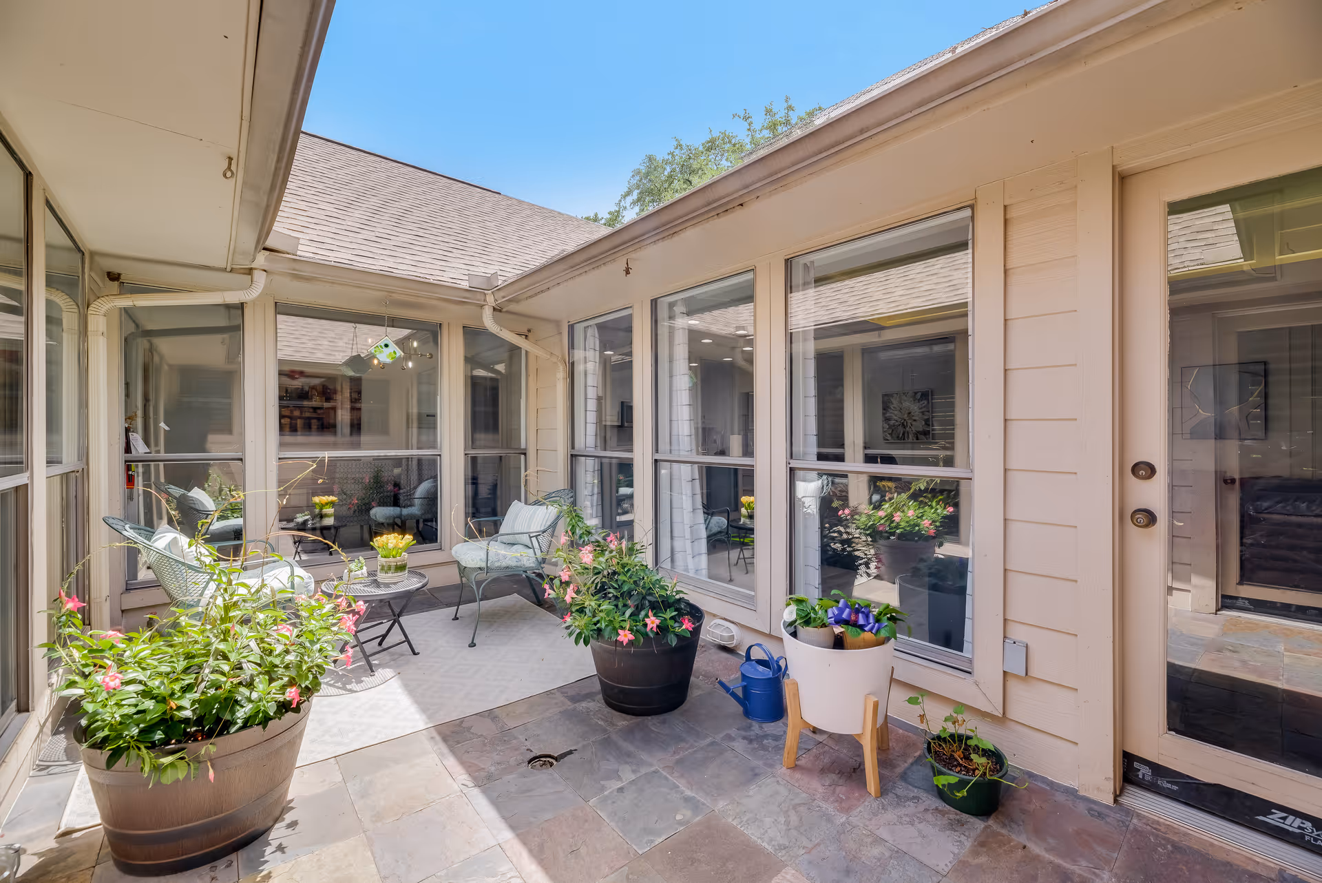 A bright and cozy outdoor patio area at New Horizon Homes featuring several potted plants with flowers, two metal chairs with cushions, a small round table with a flower pot, and large windows looking into the interior rooms. The patio is paved with stone tiles and has a clear blue sky overhead.