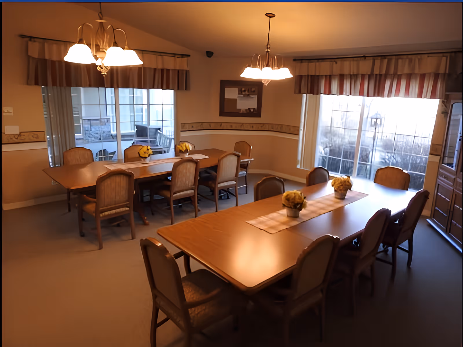 Dining room with two long wooden tables surrounded by chairs, hanging light fixtures, windows with valances, and small flower centerpieces.