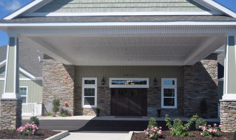 Front exterior view of Vicinia Gardens Luxury Retirement Living building entrance with stone and green siding, double dark wooden doors, two windows on either side, and a covered driveway with flower beds in front.
