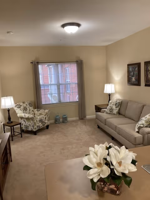 A cozy living room with beige walls and carpet. The room features a beige sofa with patterned pillows, a floral armchair, two side tables with lamps, a window with beige curtains, and a table in the foreground with a vase of white flowers.