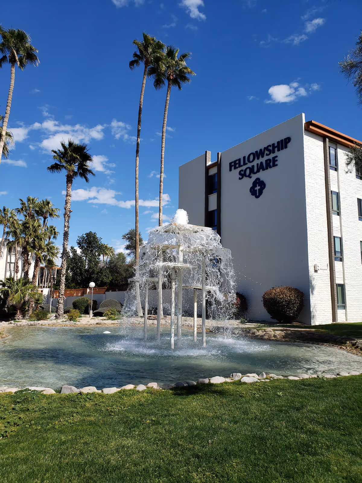 Outdoor view of Fellowship Square Tucson facility with a multi-tiered water fountain in the foreground, surrounded by a pond and green grass. Tall palm trees and other greenery are visible under a clear blue sky with a few clouds. The building has a white exterior with the name 'FELLOWSHIP SQUARE' displayed prominently on the side.