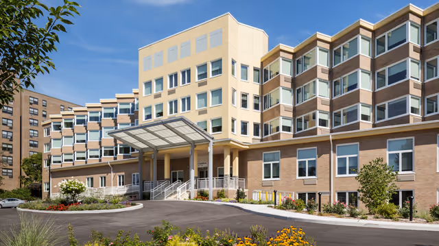 Exterior view of a multi-story senior living facility building with a covered entrance, large windows, and landscaped garden beds in front under a clear blue sky.
