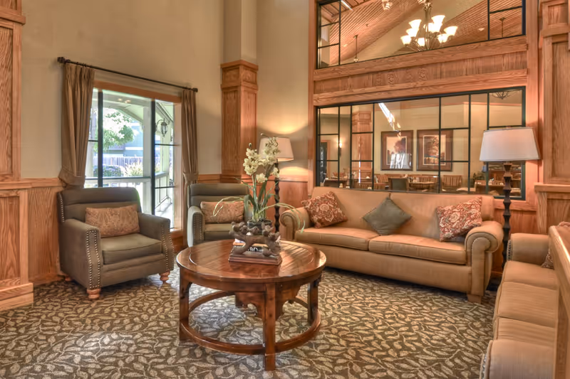 A cozy living room area with a beige sofa, two armchairs, a round wooden coffee table with a floral centerpiece, and a large window with brown curtains. The room features wood paneling, a patterned carpet, and a large mirror reflecting a dining area with framed artwork and a chandelier.