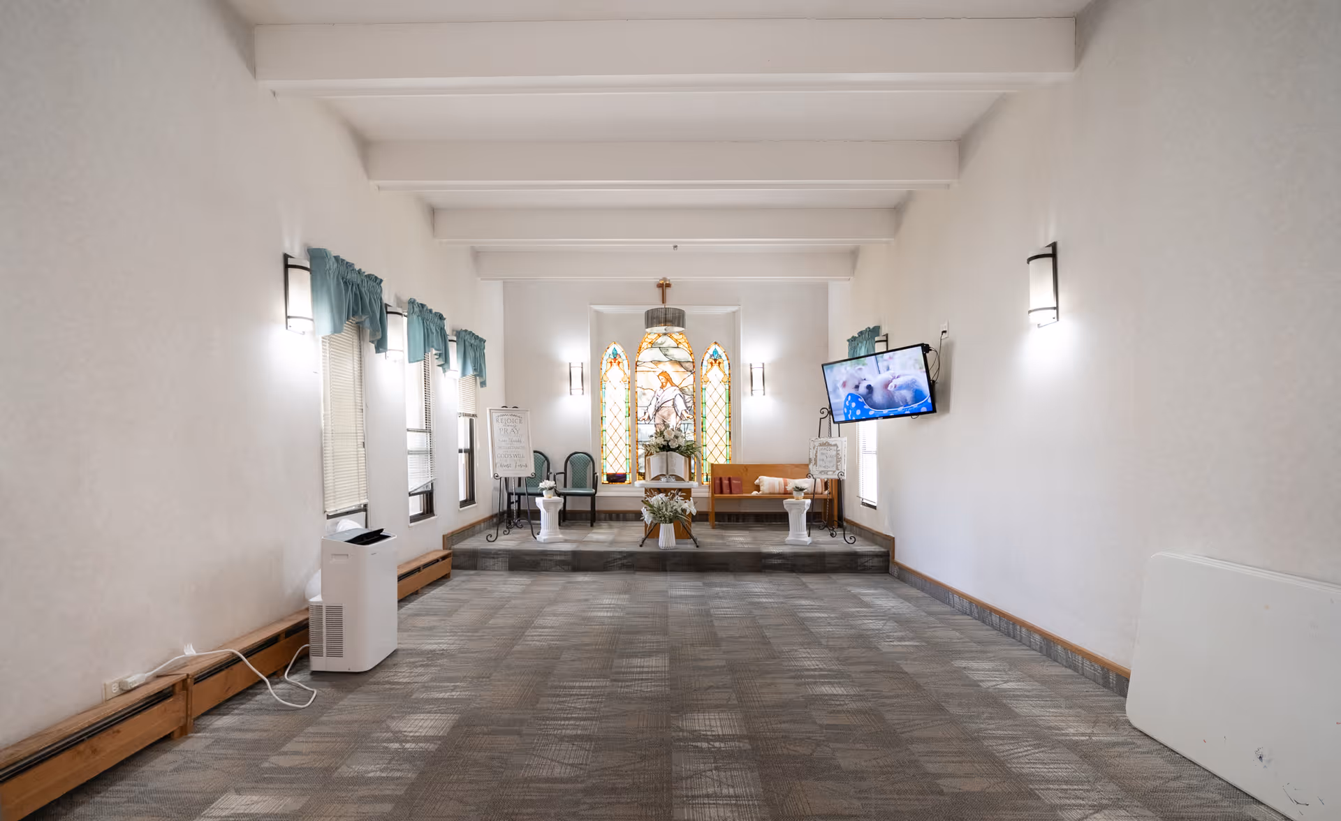 A small chapel or meditation room with a raised platform at the far end featuring a wooden bench, two green chairs, floral arrangements, and a stained glass window with religious imagery. The room has white walls, several windows with teal curtains, wall-mounted lights, a flat-screen TV on the right wall, and a patterned carpeted floor.