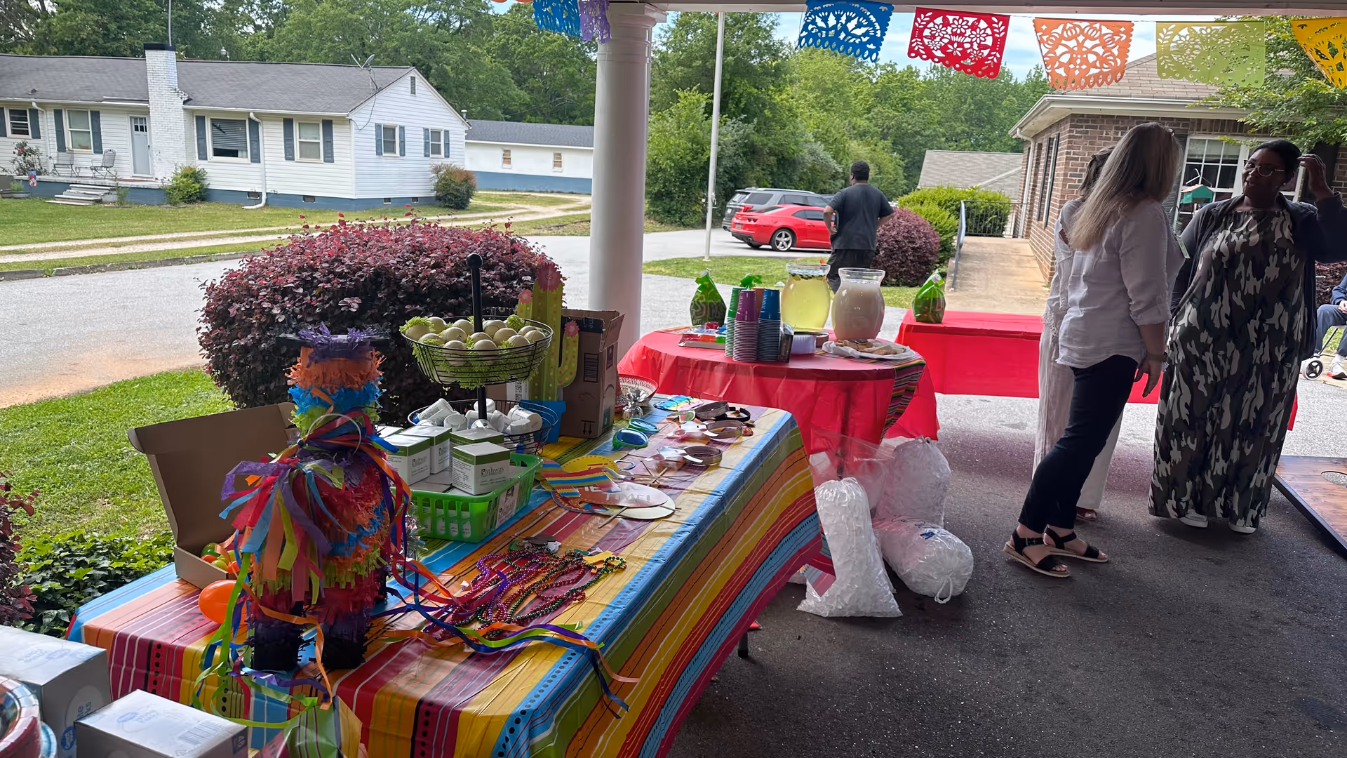 Decorated outdoor table with a colorful tablecloth holding party supplies and snacks under a covered porch while people chat nearby.