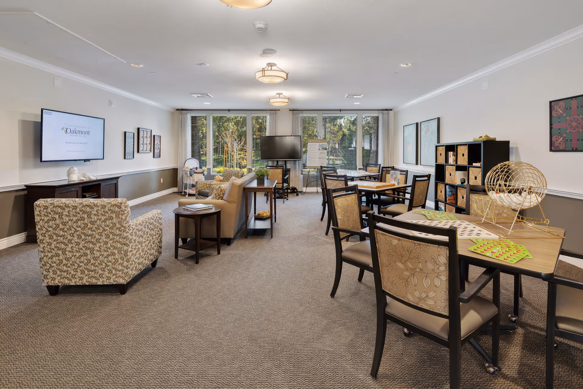 A spacious, well-lit common room with comfortable seating including armchairs and a sofa, several tables and chairs arranged for activities, a television mounted on the wall, large windows with a view of greenery outside, and decorative artwork on the walls.