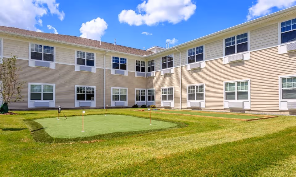 Outdoor courtyard area of a senior living facility with a small putting green and bocce ball court surrounded by a two-story beige building under a blue sky with scattered clouds.