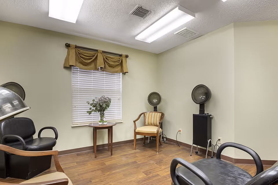 Interior room with three vintage hair dryers, two black salon chairs, one wooden chair with a cushion, a small round wooden table with a vase of flowers, and a window with blinds and a brown valance.
