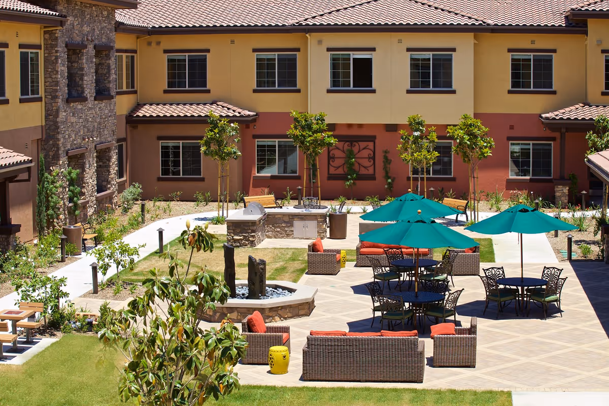 Outdoor courtyard area at Ivy Park at Escondido featuring patio seating with wicker furniture and red cushions, green umbrellas over tables, a stone fire pit, small trees, and a barbecue grill area. The courtyard is surrounded by a two-story building with yellow and brown walls and multiple windows.