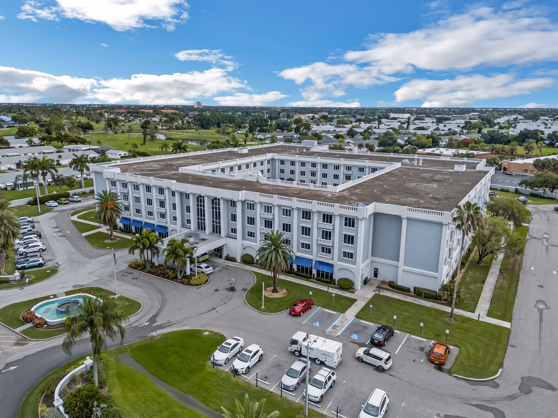 Aerial view of a large four-story white senior living building with a central courtyard, surrounding parking lot, palm trees, and landscaped grounds.