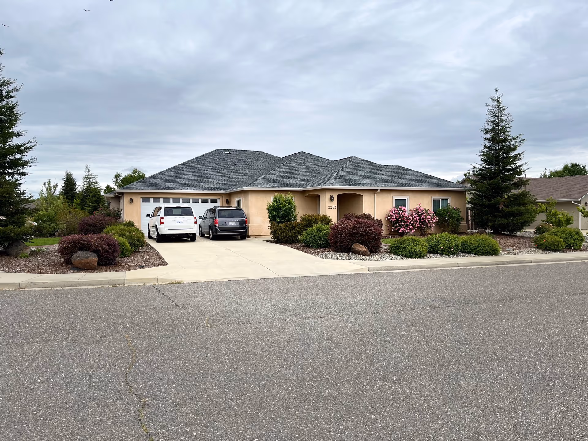 Single-story stucco house with a three-car garage, two cars parked in the driveway, and a landscaped front yard under a cloudy sky.