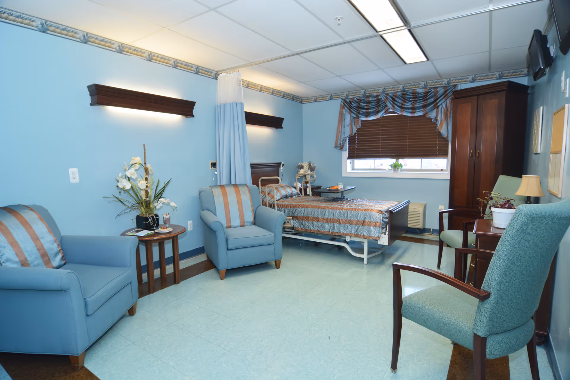 A hospital-style patient room with light blue walls and flooring. The room contains a hospital bed with striped bedding, two blue armchairs with matching striped cushions, a small round wooden table with a flower arrangement, a wooden wardrobe, a window with brown blinds and a striped valance, and a wall-mounted television.