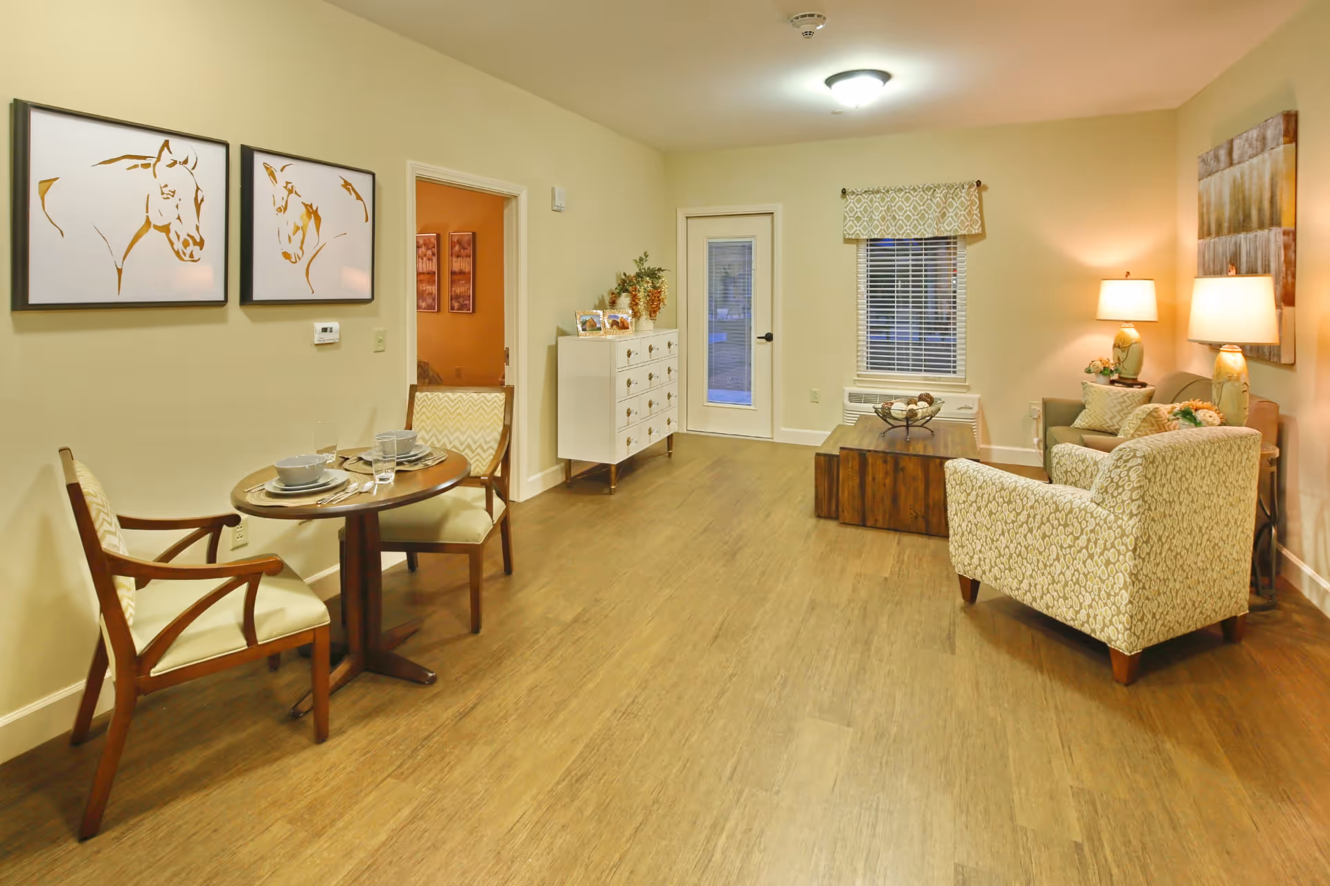 A cozy living room and dining area in a senior living facility. The room features a small round dining table set for two with chairs, two framed horse artworks on the wall, a white dresser with decorative items, a wooden coffee table, a patterned armchair, a sofa with cushions, two table lamps, and a window with blinds and a valance. The floor is wooden, and there is a door leading outside.