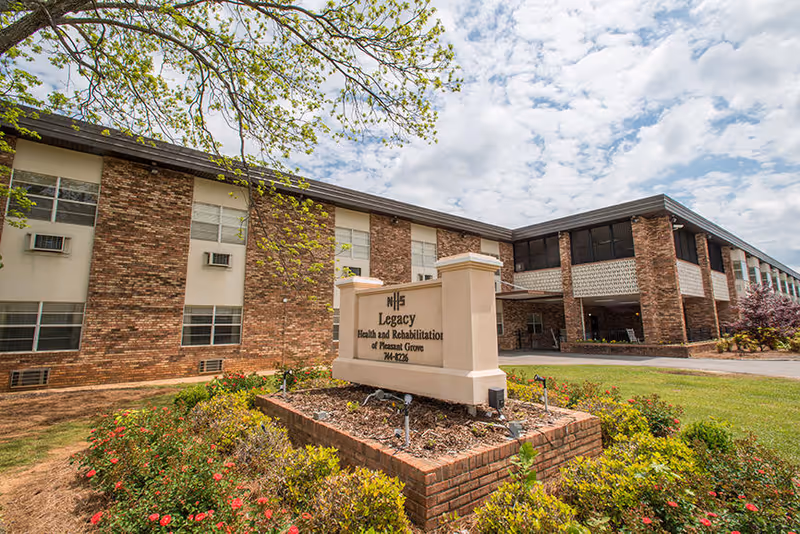 Exterior view of Legacy Health and Rehabilitation of Pleasant Grove, LLC building with a brick facade, multiple windows, and a sign in front surrounded by landscaped bushes and flowers under a partly cloudy sky.